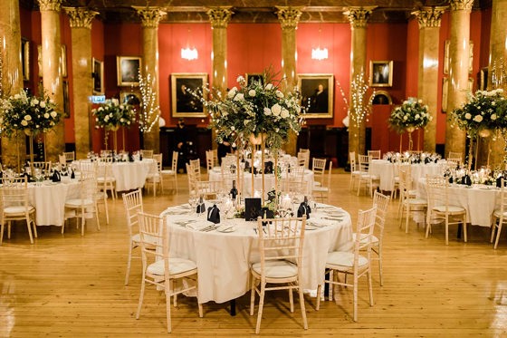 Room set up for a wedding with round tables and chairs and large floral displays 